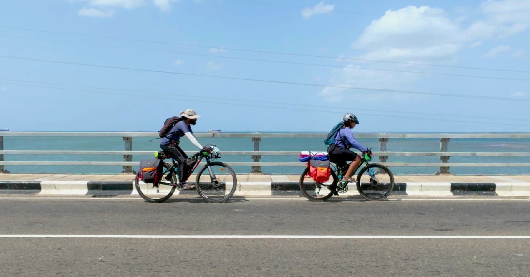 Intiaz and Subah ride loaded bicycles across a bridge beside the blue sea