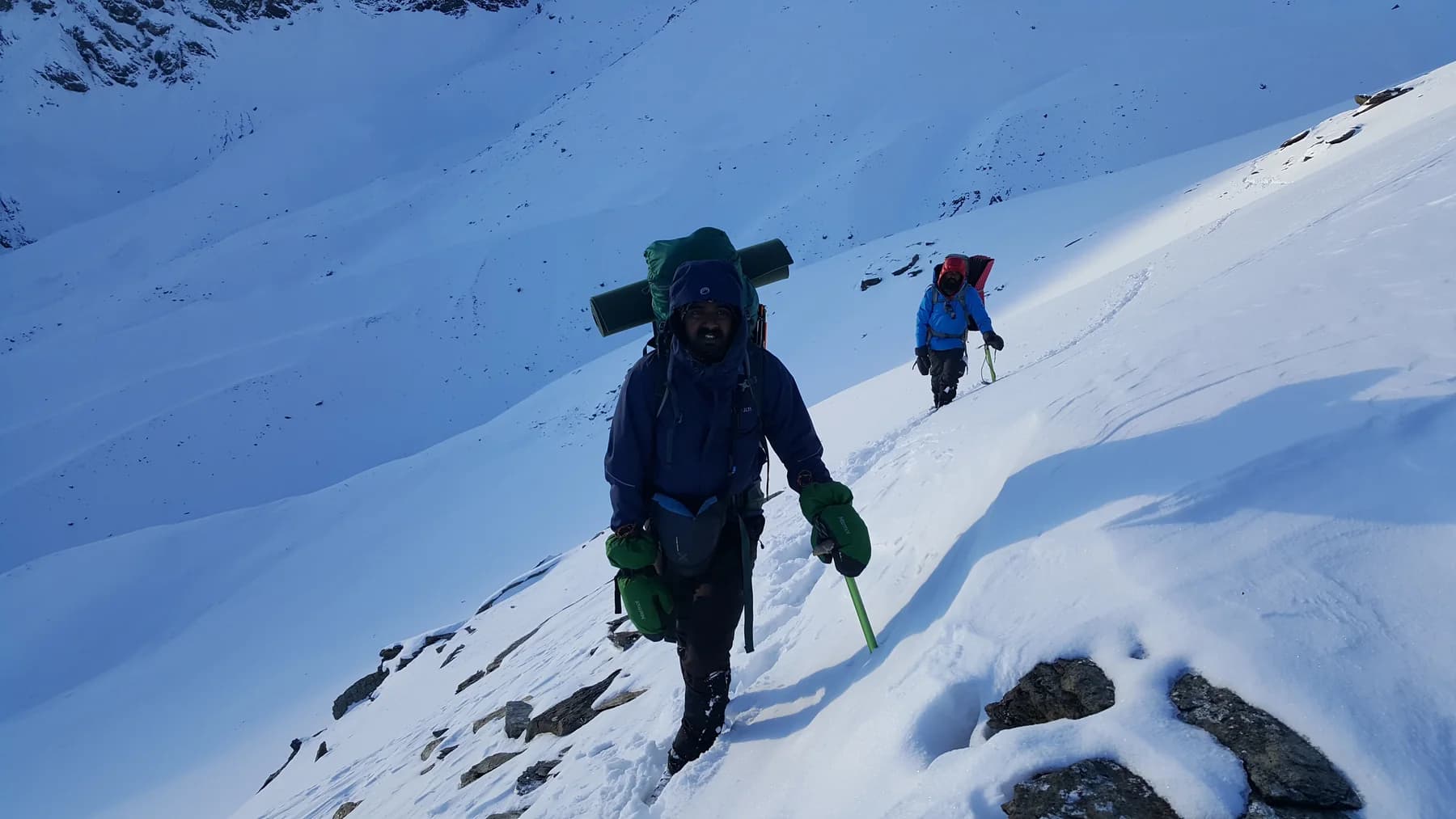 Two climbers carrying packs and ice axes across a steep snowy slope